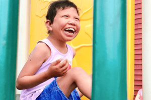 A child wearing a light purple tank top and blue shorts smiles and laughs while sitting on playground equipment, with bright yellow and red panels in the background—a joyful moment inspired by adoption from foster care. Green bars are visible in the foreground.