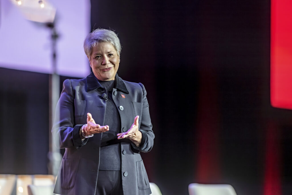 A supervisor of the year with short gray hair, wearing a dark suit, stands on stage gesturing with their hands and smiling. The background is dimly lit with soft red and purple tones.