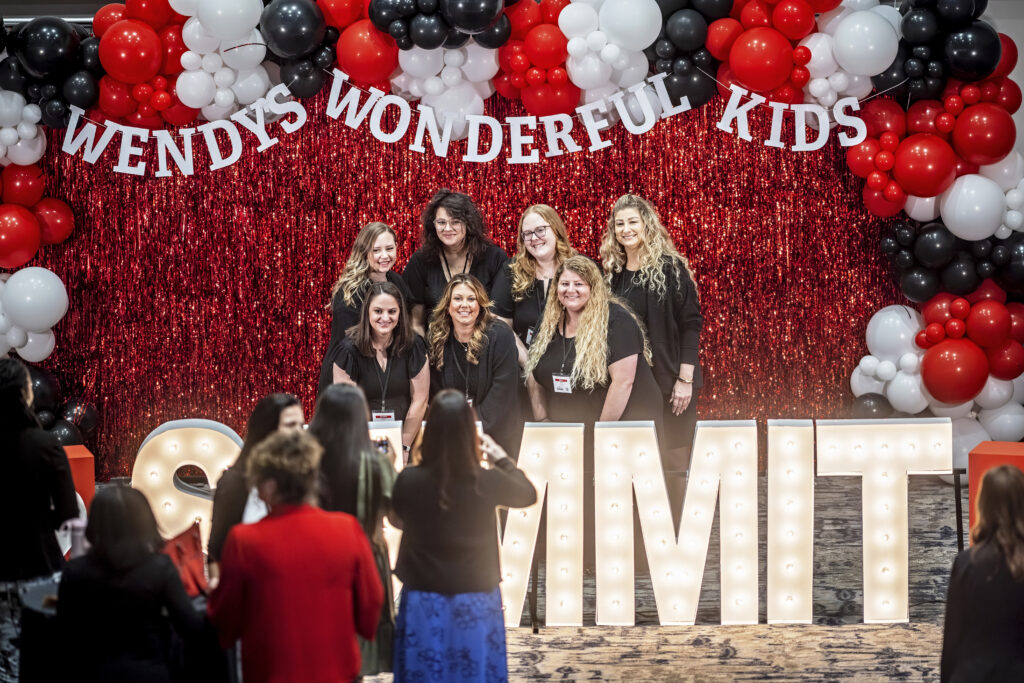 A group of smiling women, including the supervisor of the year, pose in front of large illuminated letters spelling SUMMIT, with red, black, and white balloons and a Wendys Wonderful Kids banner in the background. Audience members take photos.