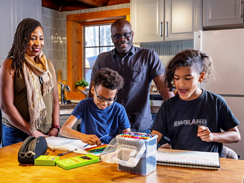 Two adults smile as two children, adopted into their family, draw and color at a kitchen table covered with art supplies. The bright, cheerful scene shows everyone focused on creative activities together.