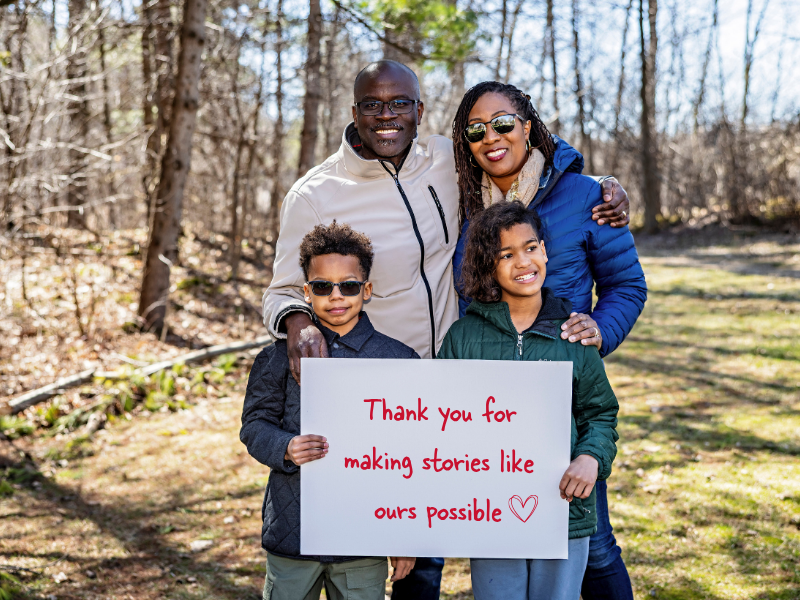 A smiling family of four stands outdoors in a wooded area, holding a sign that reads, Thank you for making adoption stories like ours possible, with a red heart drawn next to the text.