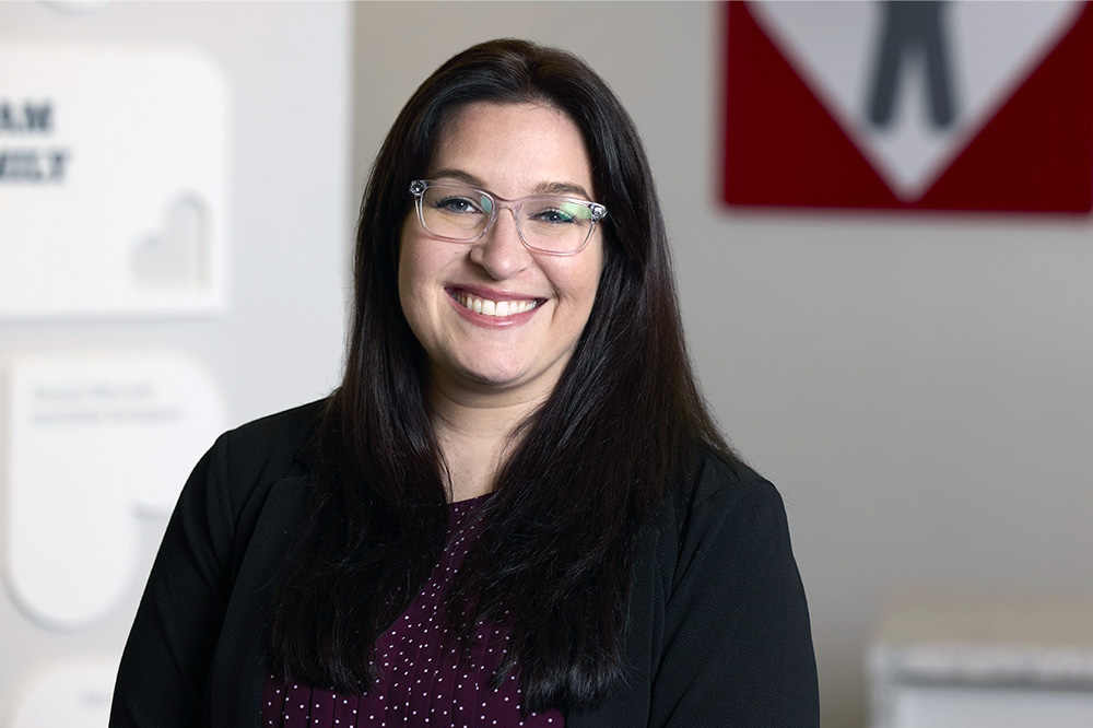 Ellen Imhoff, a woman with long dark hair, glasses, and a dark blazer, smiles at the camera indoors, with blurred office decor and a red sign in the background.